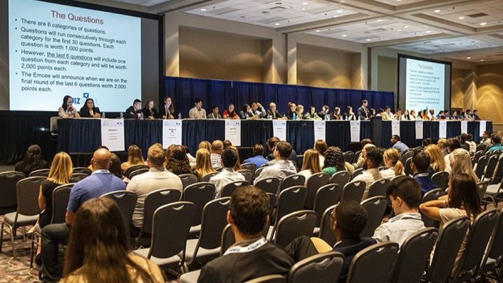 A large conference room with rows of seated participants facing a long stage. On stage, multiple individuals sit behind tables with nameplates and microphones. Two large screens display rules for the quiz bowl.